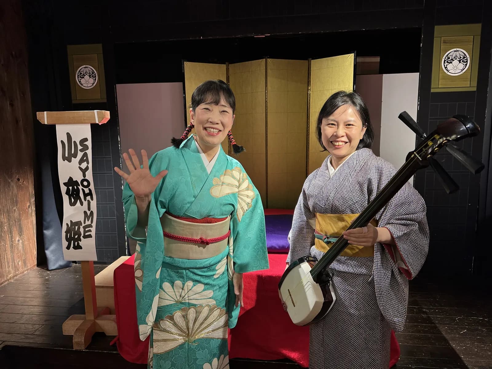 Sayohime waving beside Chiyo, who holds a shamisen, in front of a gold folding screen