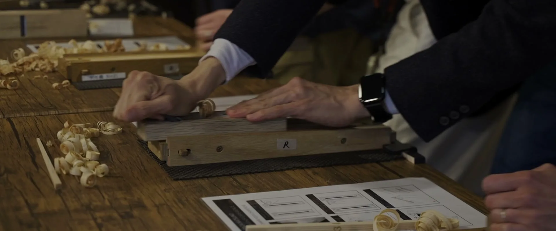 Hands feeding a strip of wood through a planing jig, shavings scattered across the bench