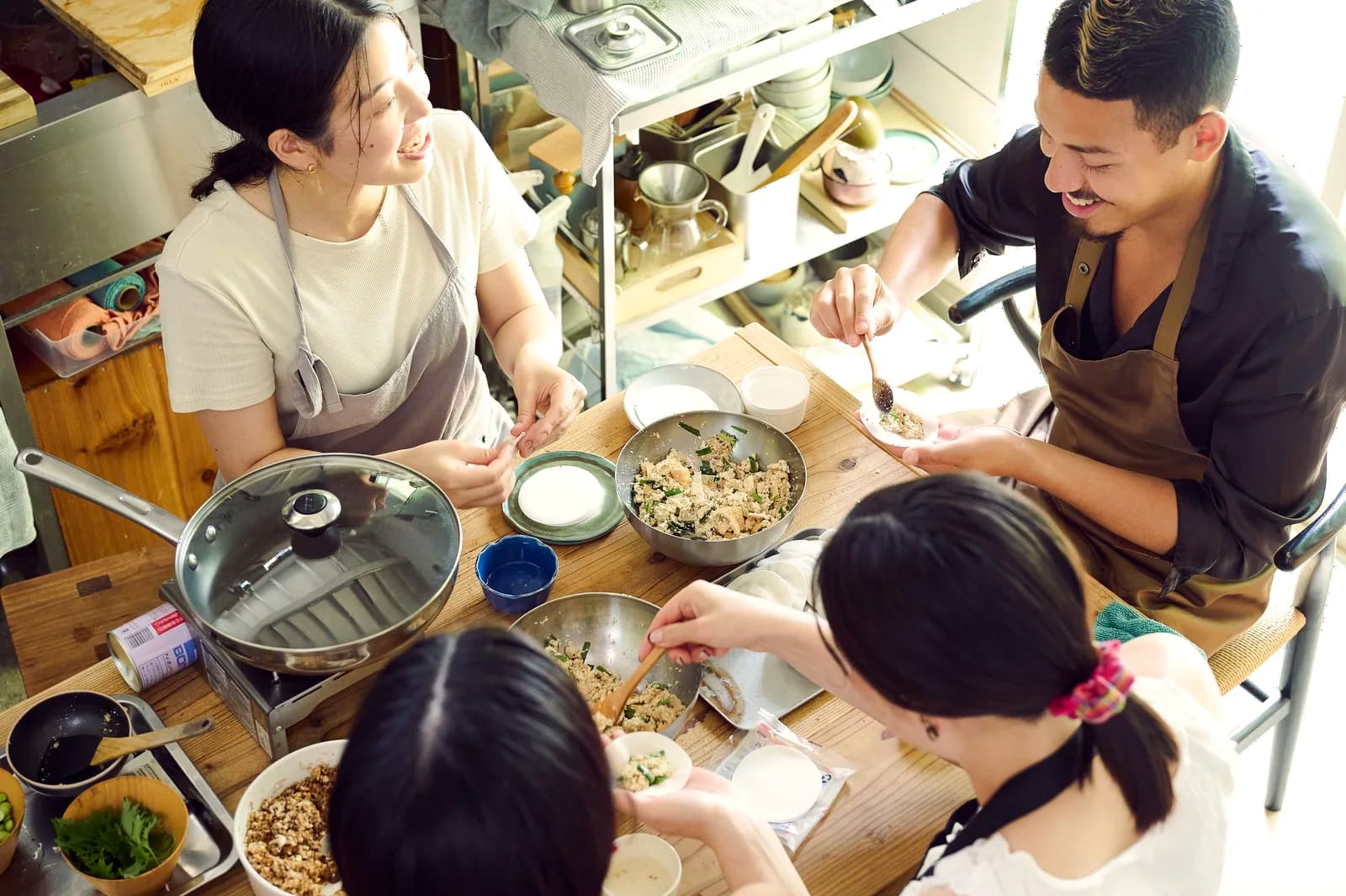 Meg, top left, and three guests around a wooden table filling gyoza wrappers, seen from above, with bowls of filling and a steamer between them