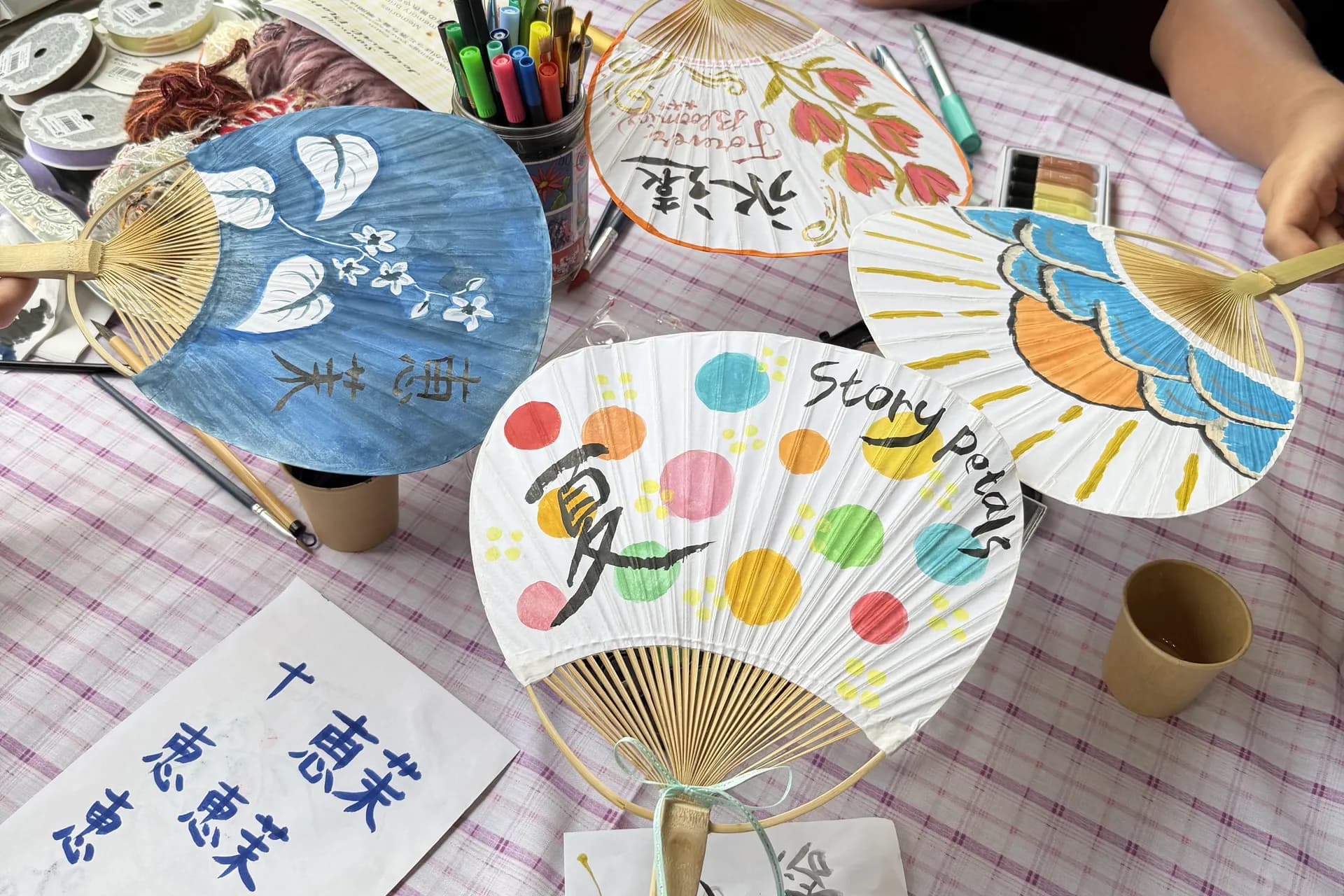Four hand-painted paper fans held over a table of paints and brushes, one lettered Story Petals