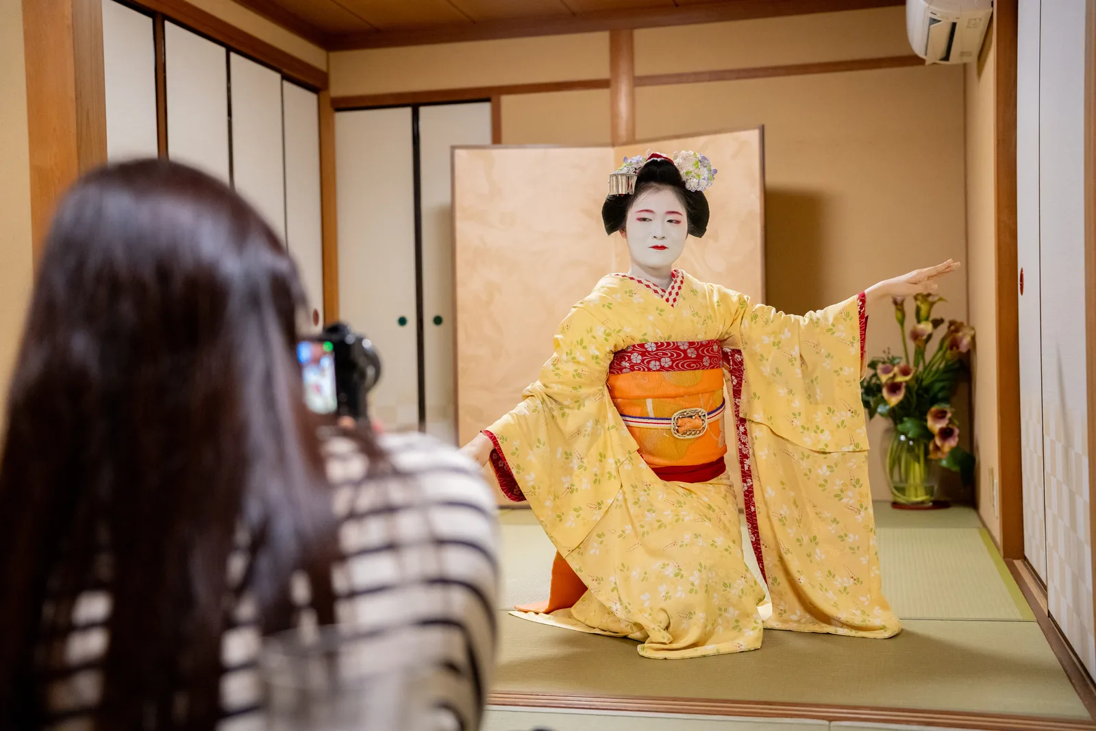 A maiko in a pale yellow kimono dancing with both arms outstretched before a gold folding screen, someone in the foreground photographing her